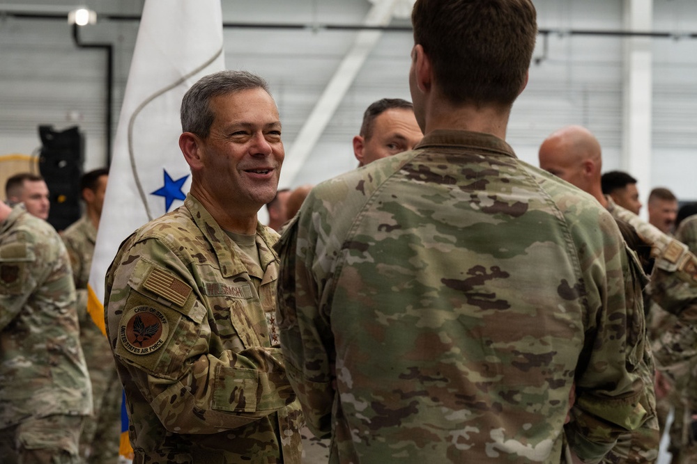 A military ceremony or gathering taking place inside what appears to be a hangar facility. A senior officer wearing a 'Chief of Staff, United States Air Force' patch and what appears to be general officer rank insignia is smiling and engaging with personnel. Multiple service members in OCP (Operational Camouflage Pattern) uniforms are present in the background.