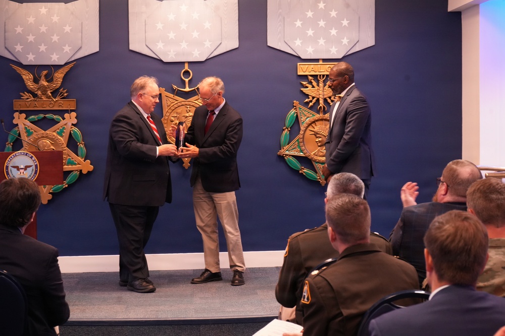 A formal award ceremony is taking place in what appears to be a U.S. Department of Defense facility, likely the Pentagon, as indicated by the Medal of Honor display emblems on the blue wall. Two men in civilian business attire are exchanging a crystal award on a raised stage, while a third man stands nearby observing. An audience of uniformed military personnel and civilians is seated in the foreground.