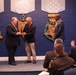 A formal award ceremony is taking place in what appears to be a U.S. Department of Defense facility, likely the Pentagon, as indicated by the Medal of Honor display emblems on the blue wall. Two men in civilian business attire are exchanging a crystal award on a raised stage, while a third man stands nearby observing. An audience of uniformed military personnel and civilians is seated in the foreground.