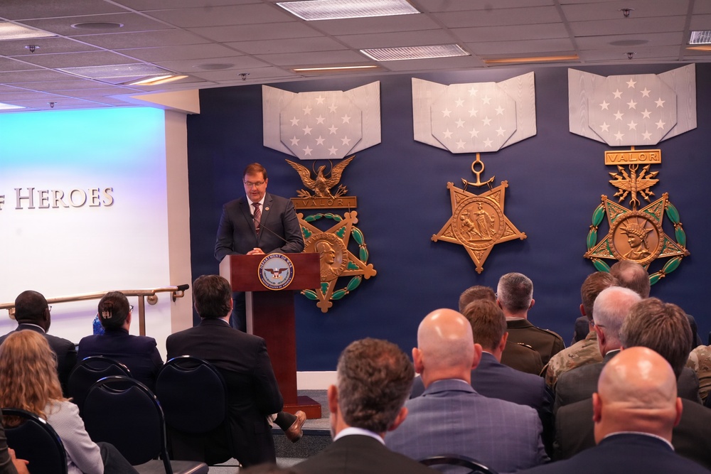 A speaker addresses an audience from a podium bearing the U.S. Department of Defense seal, in a room decorated with large representations of the three versions of the Medal of Honor (Army, Navy, and Air Force). The partial text 'HEROES' is visible on a projection screen to the left, suggesting a ceremony or event honoring military valor. The audience includes a mix of civilians in suits and military personnel in uniform.
