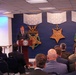 A speaker addresses an audience from a podium bearing the U.S. Department of Defense seal, in a room decorated with large representations of the three versions of the Medal of Honor (Army, Navy, and Air Force). The partial text 'HEROES' is visible on a projection screen to the left, suggesting a ceremony or event honoring military valor. The audience includes a mix of civilians in suits and military personnel in uniform.