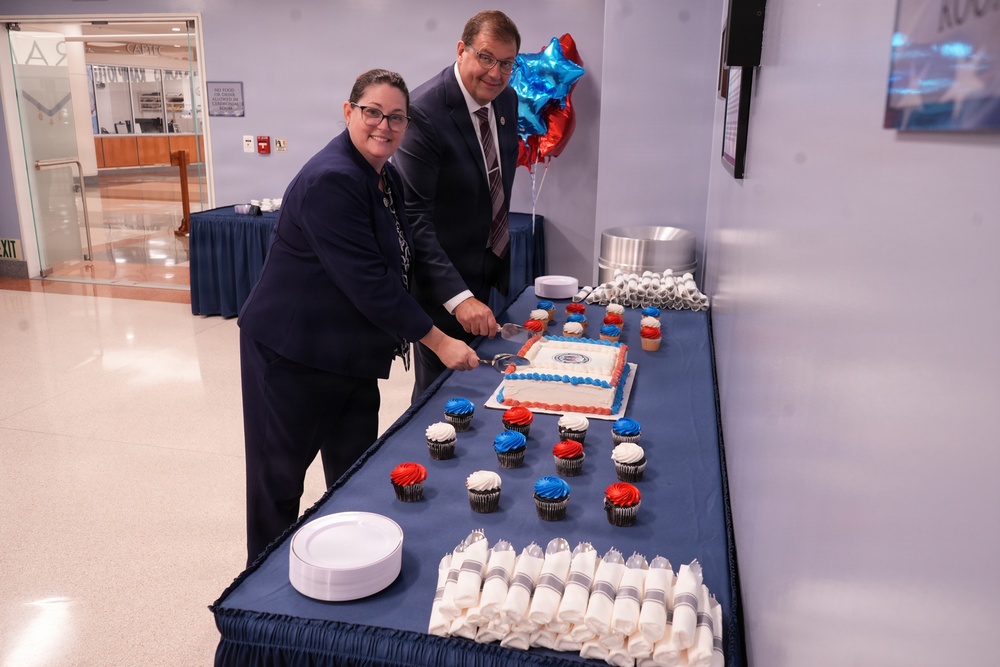 Two formally dressed individuals are cutting a decorated sheet cake at what appears to be an official ceremony or celebration. The table is adorned with red, white, and blue cupcakes and decorations, suggesting a patriotic or institutional event. Mylar balloons in red, white, and blue are visible in the background.
