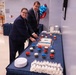 Two formally dressed individuals are cutting a decorated sheet cake at what appears to be an official ceremony or celebration. The table is adorned with red, white, and blue cupcakes and decorations, suggesting a patriotic or institutional event. Mylar balloons in red, white, and blue are visible in the background.
