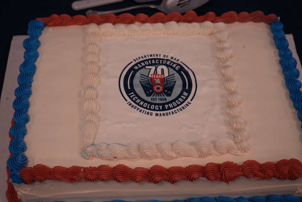 A rectangular sheet cake decorated in red, white, and blue frosting to celebrate the 70th anniversary of the Department of War Manufacturing Technology Program. The cake features a printed edible logo in the center displaying '70 Years' with the establishment date of 1956 and the tagline 'Innovating Manufacturing'. The patriotic color scheme and formal logo suggest this is an official commemorative event.