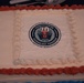 A rectangular sheet cake decorated in red, white, and blue frosting to celebrate the 70th anniversary of the Department of War Manufacturing Technology Program. The cake features a printed edible logo in the center displaying '70 Years' with the establishment date of 1956 and the tagline 'Innovating Manufacturing'. The patriotic color scheme and formal logo suggest this is an official commemorative event.