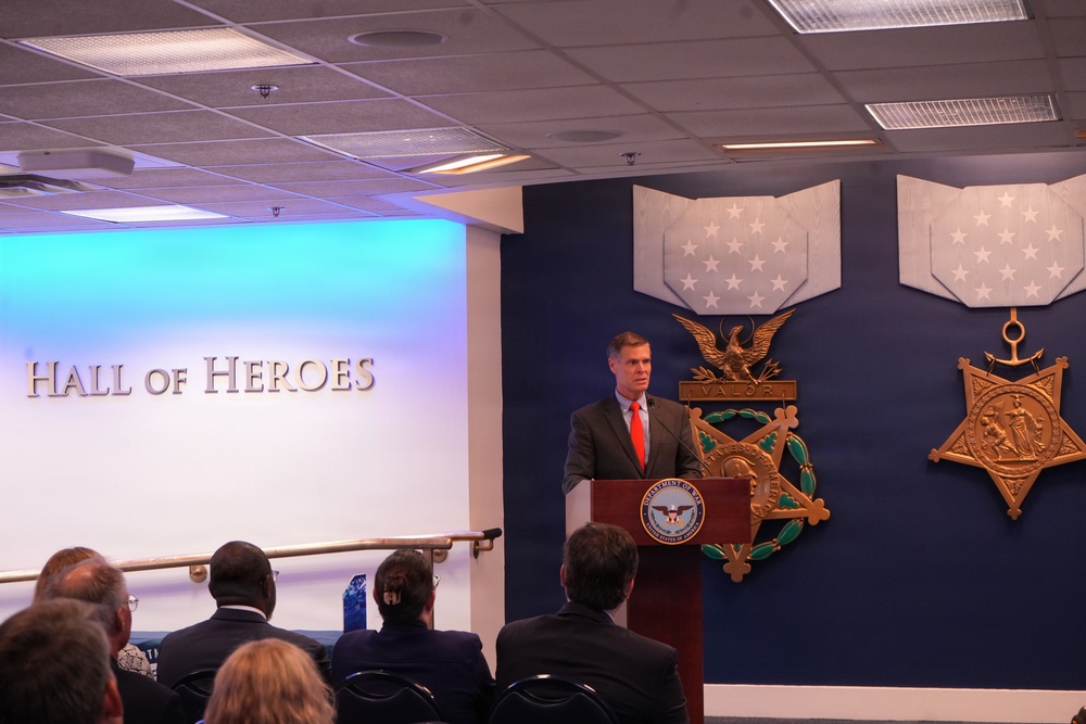 A man in a dark suit with a red tie is speaking at a podium bearing the Department of Defense seal, located in the Pentagon's Hall of Heroes. The backdrop features large decorative Medal of Honor insignia mounted on a dark blue wall. An audience is seated facing the speaker in what appears to be an official ceremony or presentation.