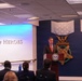 A man in a dark suit with a red tie is speaking at a podium bearing the Department of Defense seal, located in the Pentagon's Hall of Heroes. The backdrop features large decorative Medal of Honor insignia mounted on a dark blue wall. An audience is seated facing the speaker in what appears to be an official ceremony or presentation.