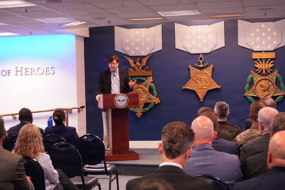 A civilian speaker addresses an audience from a podium bearing the Department of Defense seal, in a room decorated with large Medal of Honor replicas on a navy blue wall. The setting appears to be an official Pentagon or DoD facility, likely during a formal ceremony or briefing. The text 'OF HEROES' is partially visible on the left wall, suggesting this is the Pentagon's Hall of Heroes.