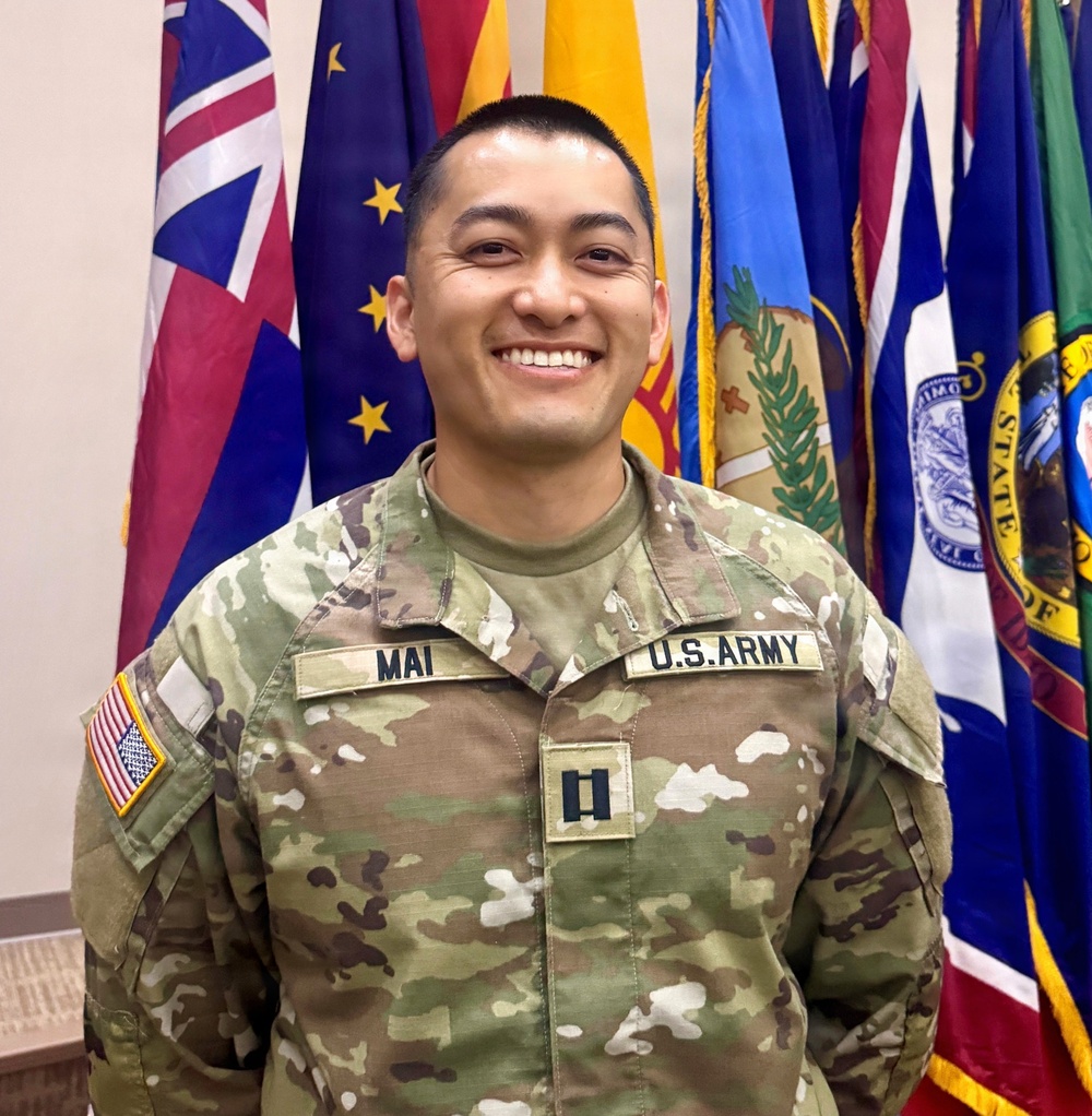 A U.S. Army Captain named Mai is posing for a formal portrait in Army Combat Uniform (ACU/OCP pattern). He is smiling broadly in front of a display of multiple U.S. state and territory flags. His rank insignia (Captain's double bars) and name tape are clearly visible on his uniform.
