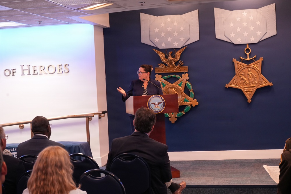 A woman is speaking at a podium bearing the Department of Defense seal in what appears to be the Pentagon's Hall of Heroes. The room features large decorative Medal of Honor replicas on a navy blue wall, including Army and Navy versions. An audience of several attendees is seated facing the speaker.