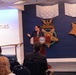 A woman is speaking at a podium bearing the Department of Defense seal in what appears to be the Pentagon's Hall of Heroes. The room features large decorative Medal of Honor replicas on a navy blue wall, including Army and Navy versions. An audience of several attendees is seated facing the speaker.