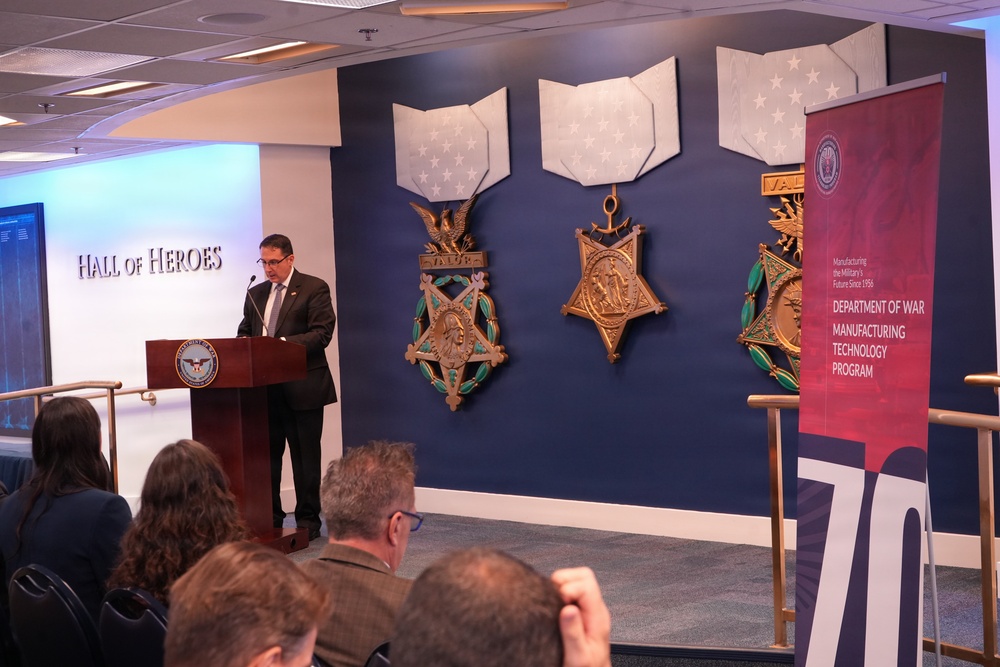 A speaker addresses an audience from a podium in the Pentagon's Hall of Heroes, standing before a display of three large Medal of Honor replicas mounted on a dark blue wall. A banner on the right identifies the event as related to the Department of War Manufacturing Technology Program, celebrating what appears to be a 70th anniversary. The audience is visible in the foreground with their backs to the camera.