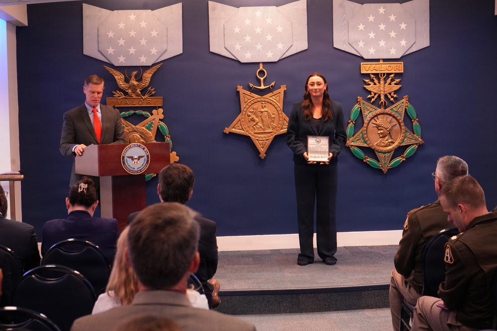 A formal award ceremony is taking place in what appears to be a U.S. Department of Defense facility, identifiable by the Department of War/Defense seal on the podium and large Medal of Honor replica emblems on the blue wall. A woman in a dark suit holds a framed award or certificate while a man in a gray suit with an orange tie stands at the podium. An audience including uniformed military personnel is seated facing the stage.
