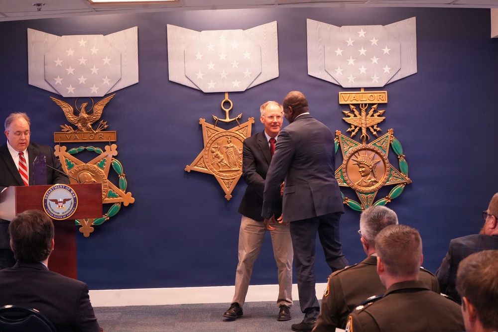 A formal ceremony is taking place in front of a display wall featuring the three branches of the Medal of Honor (Army, Navy, and Air Force versions) on a blue background. Two men in civilian attire appear to be shaking hands or exchanging an award in the center, while a speaker addresses the audience from a Department of War/Defense podium on the left. An audience including uniformed military personnel is seated in the foreground.