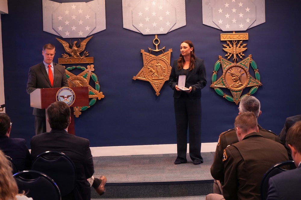 A formal military or government ceremony is taking place in what appears to be a Department of Defense facility. A woman in a dark suit stands holding what appears to be a medal or award case, while a man in a suit with a red tie speaks at a podium bearing the Department of Defense seal. Three large Medal of Honor emblems representing the Army, Navy, and Air Force branches are displayed on the blue wall behind them.
