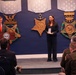 A formal military or government ceremony is taking place in what appears to be a Department of Defense facility. A woman in a dark suit stands holding what appears to be a medal or award case, while a man in a suit with a red tie speaks at a podium bearing the Department of Defense seal. Three large Medal of Honor emblems representing the Army, Navy, and Air Force branches are displayed on the blue wall behind them.