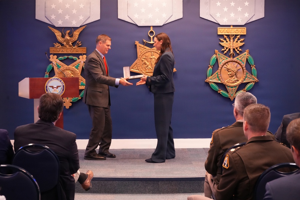 A formal award ceremony is taking place at what appears to be the Pentagon or a Department of Defense facility. A man in a gray suit with a red tie is presenting an award or plaque to a woman in a dark blazer on a raised stage. Uniformed military personnel and civilians are seated in the audience observing the presentation.
