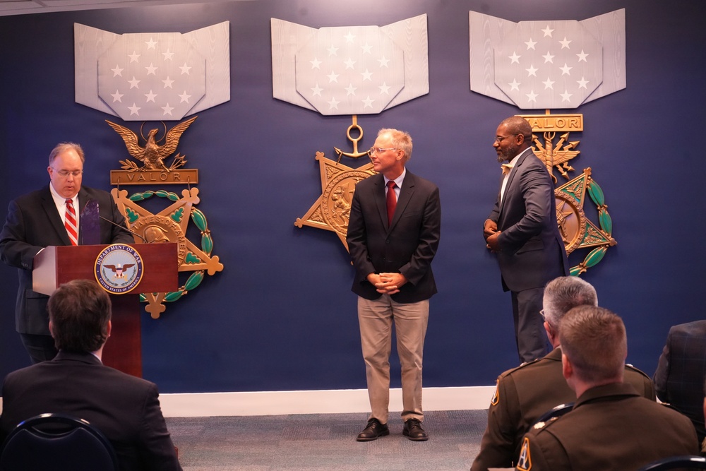 A formal ceremony is taking place in what appears to be a Department of Defense or military facility, with three large Medal of Honor replicas displayed on a blue wall behind the participants. One man stands at a podium bearing a Department of War seal while two other men stand nearby, with an audience seated in the foreground including uniformed personnel. The setting and decorations suggest an official recognition or awards ceremony.