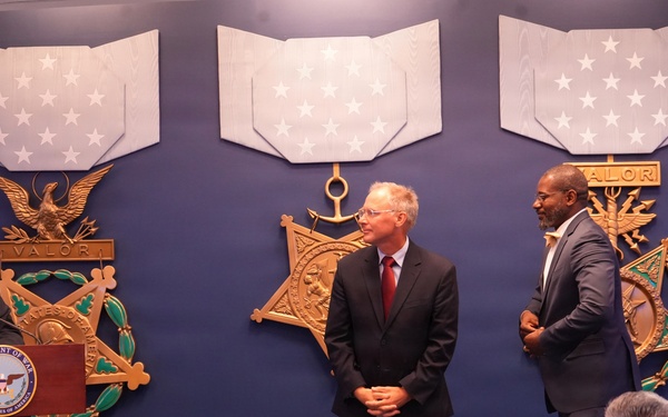 A formal ceremony is taking place in what appears to be a Department of Defense or military facility, with three large Medal of Honor replicas displayed on a blue wall behind the participants. One man stands at a podium bearing a Department of War seal while two other men stand nearby, with an audience seated in the foreground including uniformed personnel. The setting and decorations suggest an official recognition or awards ceremony.