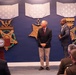 A formal ceremony is taking place in what appears to be a Department of Defense or military facility, with three large Medal of Honor replicas displayed on a blue wall behind the participants. One man stands at a podium bearing a Department of War seal while two other men stand nearby, with an audience seated in the foreground including uniformed personnel. The setting and decorations suggest an official recognition or awards ceremony.