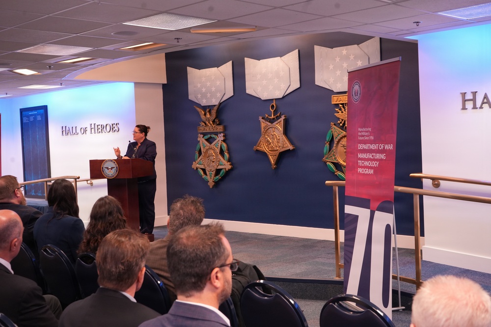 A speaker addresses an audience at a formal event held in the Pentagon's Hall of Heroes, where large Medal of Honor replicas are displayed on a navy blue wall. A red banner for the 'Department of War Manufacturing Technology Program' is visible to the right, along with a '70' anniversary marker. The audience consists of approximately 15-20 attendees seated in rows facing the podium.