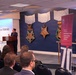 A speaker addresses an audience at a formal event held in the Pentagon's Hall of Heroes, where large Medal of Honor replicas are displayed on a navy blue wall. A red banner for the 'Department of War Manufacturing Technology Program' is visible to the right, along with a '70' anniversary marker. The audience consists of approximately 15-20 attendees seated in rows facing the podium.