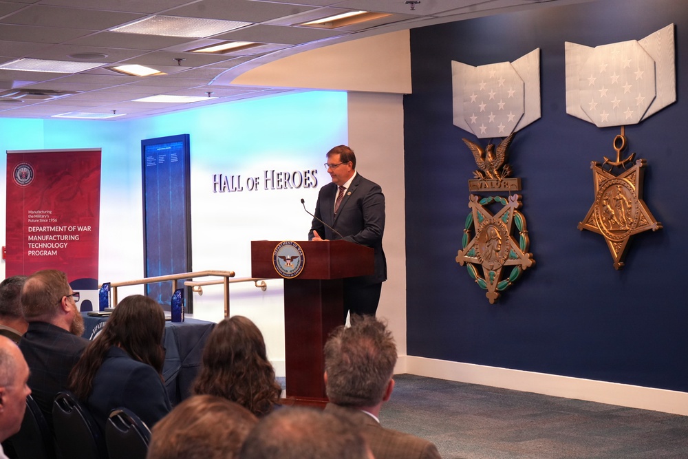 A man in a dark suit is speaking at a podium bearing the Department of Defense seal in the Pentagon's Hall of Heroes. Behind him are large decorative representations of the Medal of Honor (Army and Navy versions) mounted on a dark blue wall. An audience is seated in the foreground, and a banner for the 'Department of War Manufacturing Technology Program' is visible to the left.