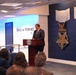A man in a dark suit is speaking at a podium bearing the Department of Defense seal in the Pentagon's Hall of Heroes. Behind him are large decorative representations of the Medal of Honor (Army and Navy versions) mounted on a dark blue wall. An audience is seated in the foreground, and a banner for the 'Department of War Manufacturing Technology Program' is visible to the left.