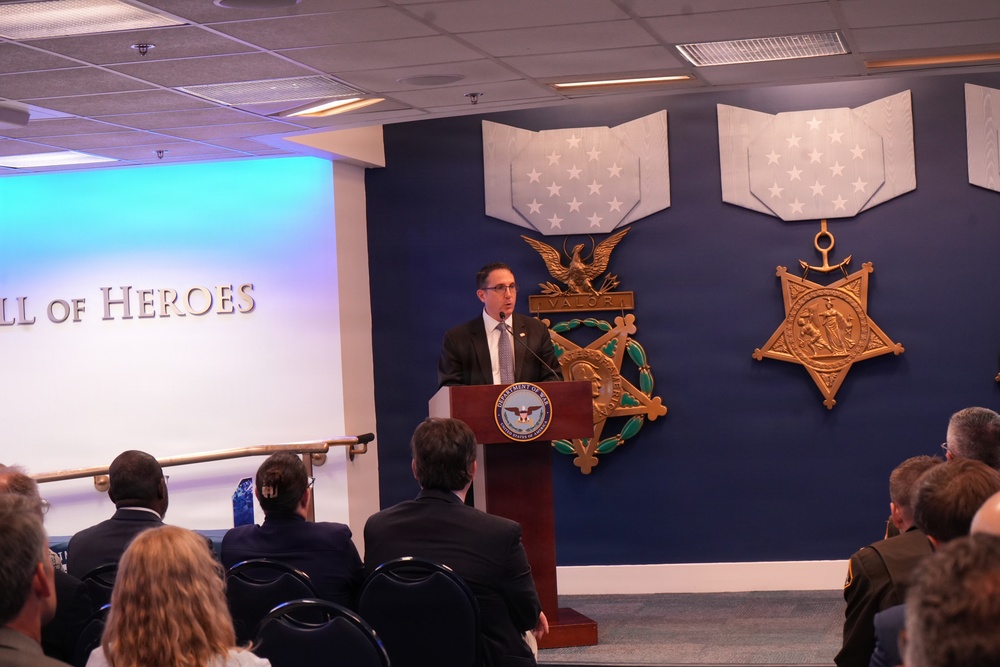 A man in a dark suit is speaking at a podium bearing the Department of Defense seal, located in what appears to be the Pentagon's Hall of Heroes. The blue wall behind him displays large decorative Medal of Honor replicas, including the Army and Navy versions. An audience of attendees is seated facing the speaker during what appears to be an official ceremony or presentation.