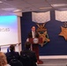 A man in a dark suit is speaking at a podium bearing the Department of Defense seal, located in what appears to be the Pentagon's Hall of Heroes. The blue wall behind him displays large decorative Medal of Honor replicas, including the Army and Navy versions. An audience of attendees is seated facing the speaker during what appears to be an official ceremony or presentation.