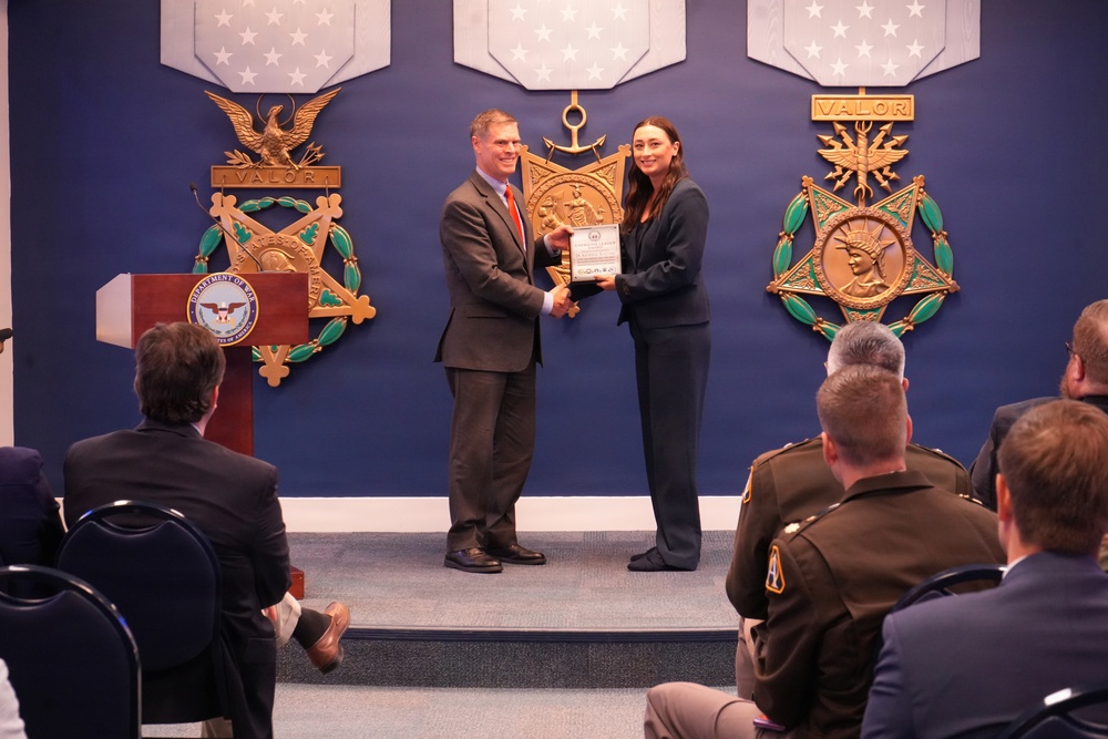A formal award ceremony is taking place at what appears to be a U.S. Department of Defense facility, identifiable by the podium seal and Medal of Honor decorations on the wall. A man in a gray suit with a red tie is presenting a plaque or certificate to a woman in a dark blazer on a small stage. An audience including military personnel in uniform and civilians observe the presentation.