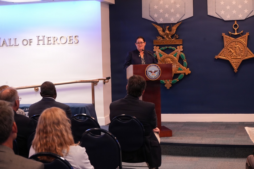 A woman in a dark blazer is speaking at a podium bearing the Department of Defense seal in the Pentagon's Hall of Heroes. The room features large Medal of Honor replicas mounted on a dark blue wall, and an audience seated in chairs faces the speaker. The setting suggests an official government ceremony or briefing.