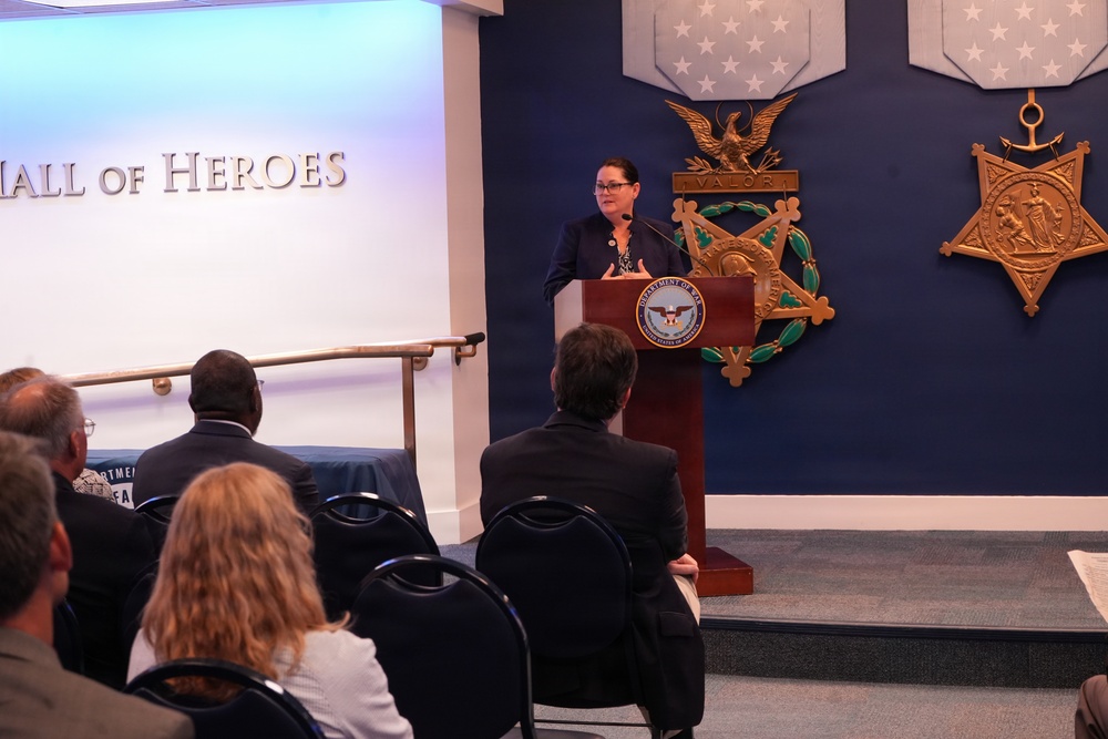 A woman is speaking at a podium bearing the Department of Defense seal in the Pentagon's Hall of Heroes. The room features large Medal of Honor replicas on a dark blue wall, and an audience of approximately 8-10 people is seated facing the speaker. The setting suggests an official ceremony or formal presentation.