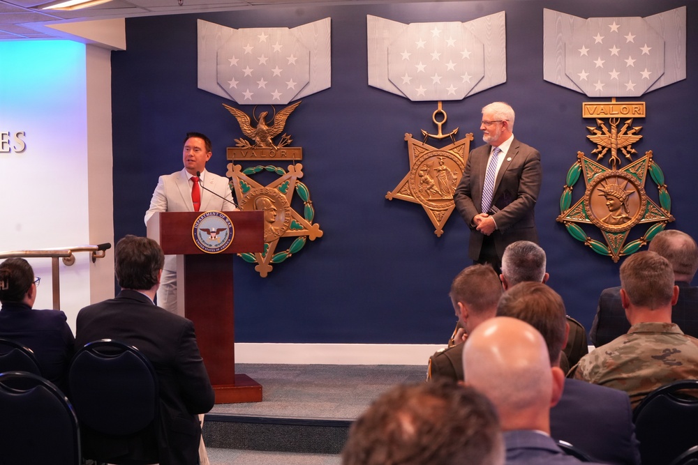 A formal ceremony is taking place at what appears to be the Pentagon or a Department of Defense facility, with a speaker at a podium bearing the Department of Defense seal. Three large Medal of Honor replicas are displayed on a blue wall behind the speakers, including Army, Navy, and Air Force versions. An audience of military personnel and civilians is seated facing the podium.