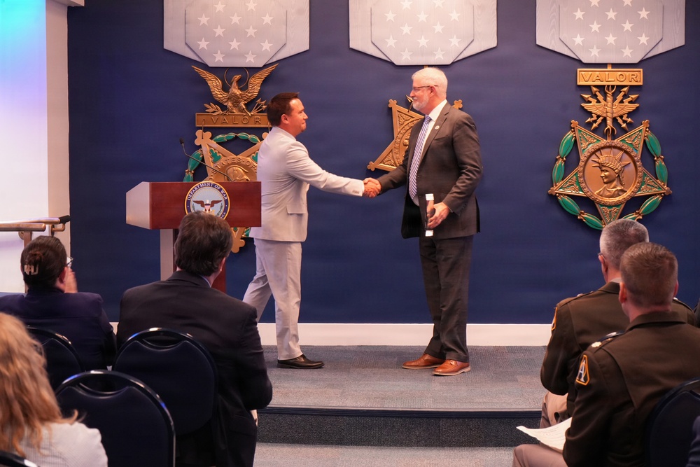 Two men in civilian attire are shaking hands on a stage at what appears to be an official U.S. Department of Defense ceremony. The setting features prominent Medal of Honor emblems and 'VALOR' insignia on a blue wall backdrop, along with a Department of Defense podium. An audience of attendees, including uniformed military personnel, is seated in the foreground.