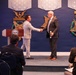 Two men in civilian attire are shaking hands on a stage at what appears to be an official U.S. Department of Defense ceremony. The setting features prominent Medal of Honor emblems and 'VALOR' insignia on a blue wall backdrop, along with a Department of Defense podium. An audience of attendees, including uniformed military personnel, is seated in the foreground.