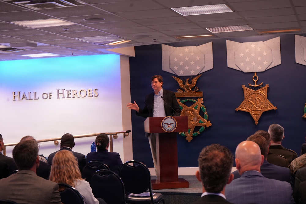 A man in business casual attire is speaking at a podium bearing the Department of Defense seal in the Pentagon's Hall of Heroes. The room is filled with an audience seated in chairs, listening to the presentation. The backdrop features large decorative Medal of Honor replicas mounted on a blue wall.