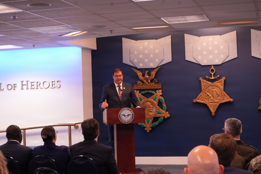 A man in a dark suit is speaking at a podium bearing the Department of Defense seal, positioned in front of a blue wall displaying large decorative Medal of Honor replicas. The text 'of Heroes' is visible on a projection screen to the left, suggesting this is a 'Hall of Heroes' ceremony or event. An audience is seated facing the speaker in what appears to be an official Pentagon or DoD facility.