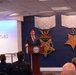 A man in a dark suit is speaking at a podium bearing the Department of Defense seal, positioned in front of a blue wall displaying large decorative Medal of Honor replicas. The text 'of Heroes' is visible on a projection screen to the left, suggesting this is a 'Hall of Heroes' ceremony or event. An audience is seated facing the speaker in what appears to be an official Pentagon or DoD facility.