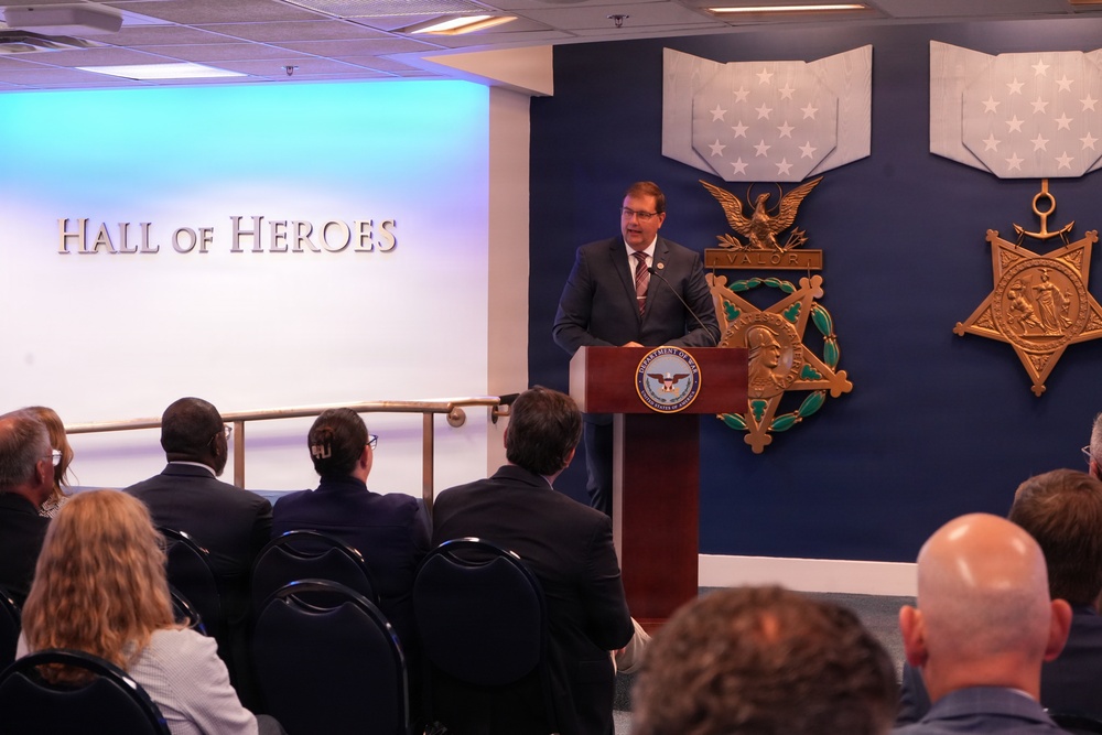 A man in a dark suit is speaking at a podium bearing the Department of Defense seal in the Hall of Heroes, located in the Pentagon. The background features large decorative representations of the Medal of Honor, including the Army and Navy versions. An audience of approximately 15-20 people is seated facing the speaker.