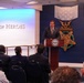 A man in a dark suit is speaking at a podium bearing the Department of Defense seal in the Hall of Heroes, located in the Pentagon. The background features large decorative representations of the Medal of Honor, including the Army and Navy versions. An audience of approximately 15-20 people is seated facing the speaker.