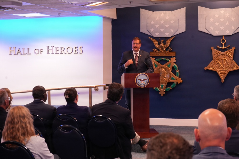 A man in a dark suit is delivering a speech at a podium bearing the Department of Defense seal, located in the Pentagon's Hall of Heroes. The backdrop features large Medal of Honor emblems on a navy blue wall, including the Army and Navy versions of the medal. An audience of approximately 15-20 people is seated facing the speaker.