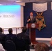 A man in a dark suit is delivering a speech at a podium bearing the Department of Defense seal, located in the Pentagon's Hall of Heroes. The backdrop features large Medal of Honor emblems on a navy blue wall, including the Army and Navy versions of the medal. An audience of approximately 15-20 people is seated facing the speaker.