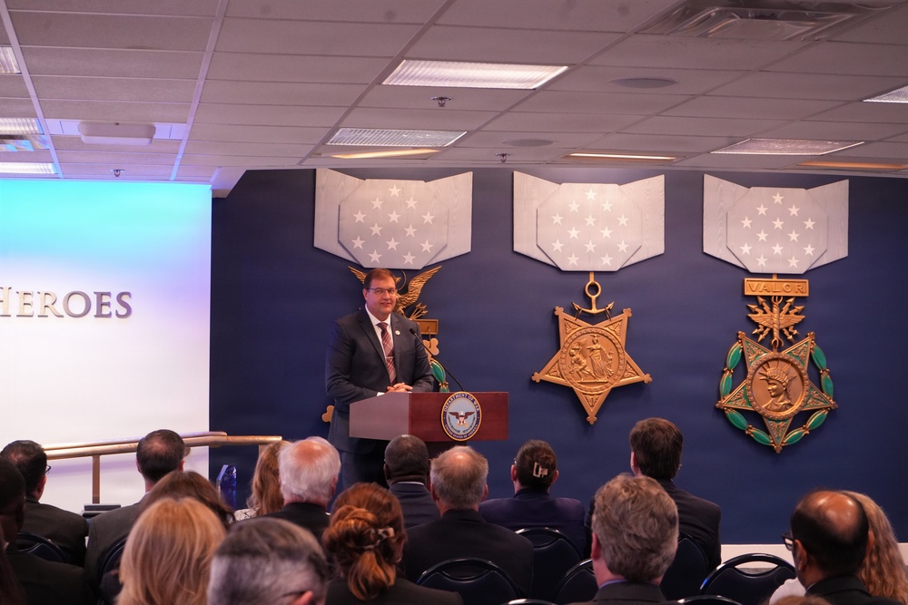 A man in a dark suit is speaking at a podium bearing what appears to be the Department of Defense seal, in front of an audience seated in rows. The backdrop features large decorative representations of the Medal of Honor variants — Army, Navy, and Air Force — mounted on a dark blue wall. A projection screen to the left displays the partial word 'HEROES', suggesting a military honors or veterans recognition event.