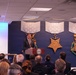 A man in a dark suit is speaking at a podium bearing what appears to be the Department of Defense seal, in front of an audience seated in rows. The backdrop features large decorative representations of the Medal of Honor variants — Army, Navy, and Air Force — mounted on a dark blue wall. A projection screen to the left displays the partial word 'HEROES', suggesting a military honors or veterans recognition event.