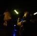 A night operations scene on what appears to be a military aircraft carrier flight deck. A deck crew member wearing a flight helmet and hearing protection is using illuminated wands (glow sticks or light wands) to signal or marshal an aircraft during nighttime operations. Blue deck lighting and the silhouette of a military aircraft are visible in the background.