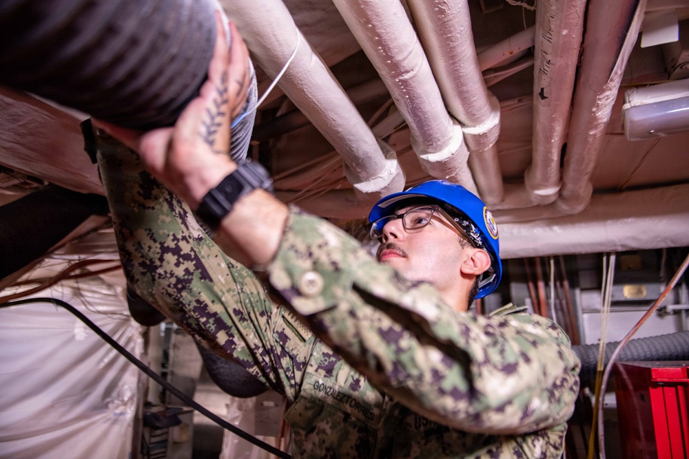 A U.S. Navy sailor in digital camouflage (NWU Type III) uniform is working on overhead piping or ductwork in what appears to be a ship's interior or industrial space. The sailor is wearing a blue hard hat with a Navy emblem and safety glasses, indicating compliance with personal protective equipment requirements. The name tape on the uniform reads 'GONZALEZTORRES'.