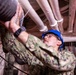 A U.S. Navy sailor in digital camouflage (NWU Type III) uniform is working on overhead piping or ductwork in what appears to be a ship's interior or industrial space. The sailor is wearing a blue hard hat with a Navy emblem and safety glasses, indicating compliance with personal protective equipment requirements. The name tape on the uniform reads 'GONZALEZTORRES'.