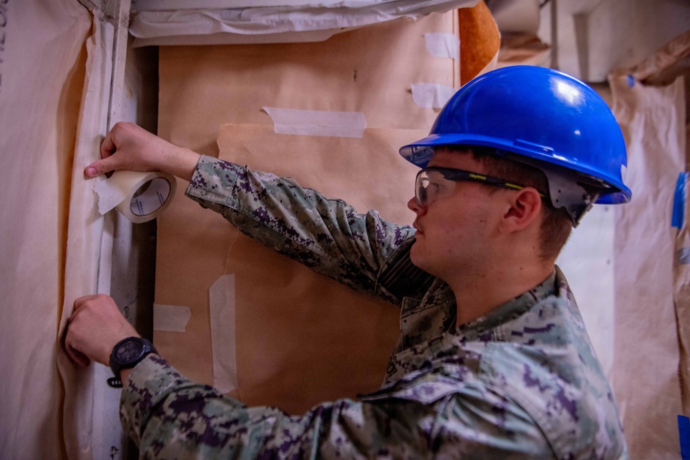 A military service member wearing U.S. Navy digital camouflage (NWU Type I) and a blue hard hat is applying masking tape to brown kraft paper secured against a fabric wall or tent surface. The individual is also wearing safety glasses and a black wristwatch, suggesting a construction or preparation task in a field or temporary facility environment. Multiple pieces of tape are already visible on the paper and surrounding surfaces.