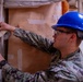 A military service member wearing U.S. Navy digital camouflage (NWU Type I) and a blue hard hat is applying masking tape to brown kraft paper secured against a fabric wall or tent surface. The individual is also wearing safety glasses and a black wristwatch, suggesting a construction or preparation task in a field or temporary facility environment. Multiple pieces of tape are already visible on the paper and surrounding surfaces.